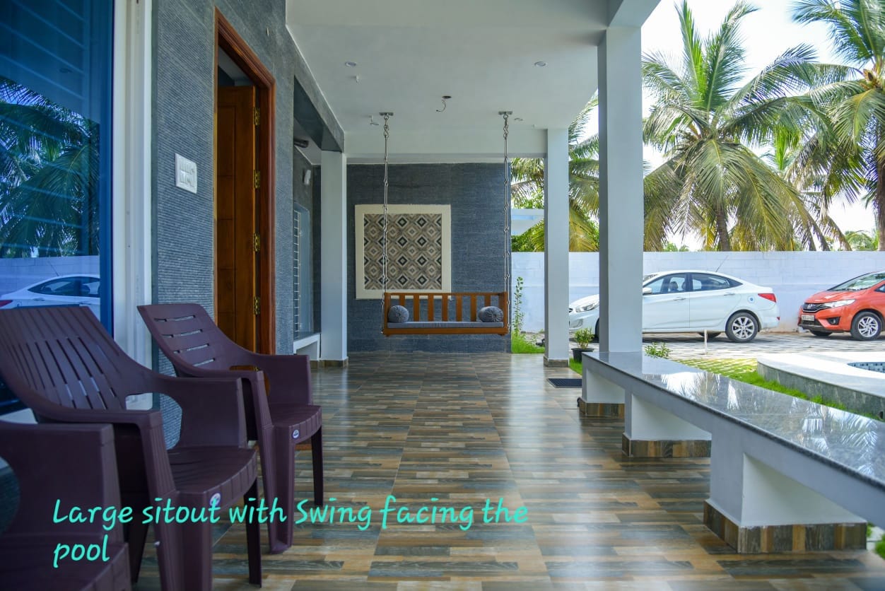 A large covered sitout area featuring a hanging wooden swing, plastic chairs, and tiled flooring.