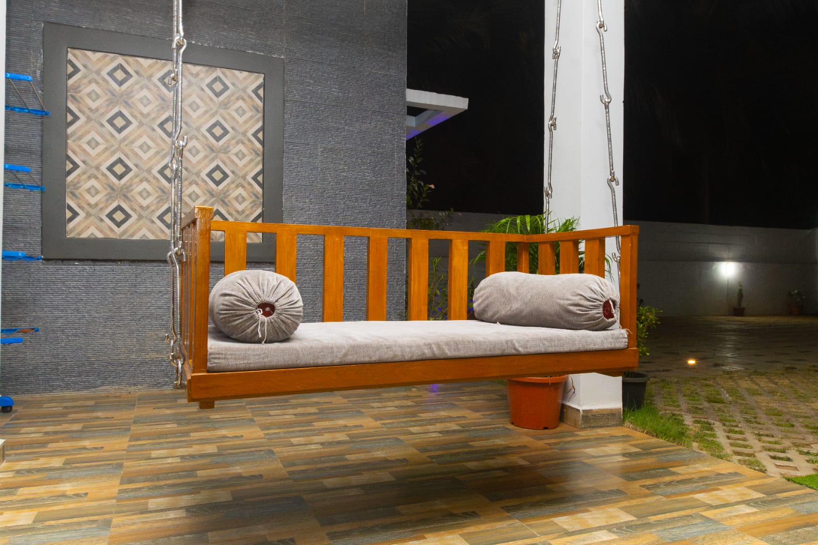 A wooden hanging swing bench with grey cushions on a tiled outdoor porch.