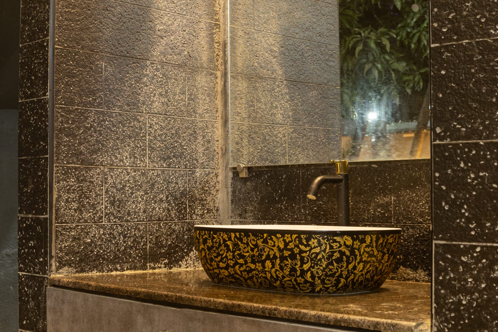 A decorative black and gold floral washbasin sitting on a countertop against dark tiled walls.